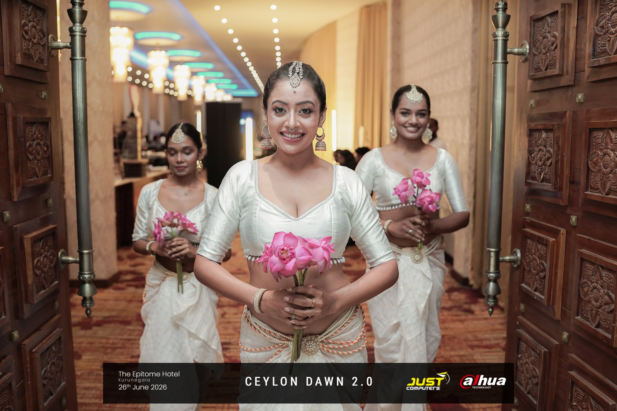 Dancers in white traditional dress carrying pink lotus flowers through carved wooden doors at The Epitome Hotel, Kurunegala